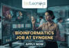 A high-tech promotional graphic for a Bioinformatics Job at Syngene, featuring the Biotecnika logo at the top. The image depicts a female scientist in a professional office setting, using a digital stylus to interact with glowing, transparent holographic screens. These displays show complex biological data, including 3D molecular protein structures, genetic heatmaps, and phylogenetic trees. In the background, a modern laboratory with other researchers is visible. The central text reads "BIOINFORMATICS JOB AT SYNGENE" in bold gold lettering, with a dark blue "APPLY NOW" button located at the bottom center.