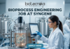 Bioprocess Engineering Job at Syngene Biopharma Analyst Role in Bangalore Apply Now! A professional featured image for a biotechnology job opening at Lupin. The scene depicts a female scientist in a modern, high-tech laboratory wearing a white lab coat and safety gear, working with a multichannel pipette and a multi-well plate. The background shows computer monitors with molecular structures and advanced lab equipment. The image includes the Biotecnika logo at the top center, the bold text "BIOTECHNOLOGY JOB AT LUPIN" in the middle, and a prominent "APPLY NOW" button at the bottom.