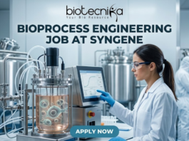 Bioprocess Engineering Job at Syngene Biopharma Analyst Role in Bangalore Apply Now! A professional featured image for a biotechnology job opening at Lupin. The scene depicts a female scientist in a modern, high-tech laboratory wearing a white lab coat and safety gear, working with a multichannel pipette and a multi-well plate. The background shows computer monitors with molecular structures and advanced lab equipment. The image includes the Biotecnika logo at the top center, the bold text "BIOTECHNOLOGY JOB AT LUPIN" in the middle, and a prominent "APPLY NOW" button at the bottom.