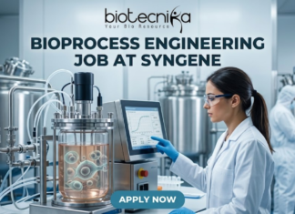 Bioprocess Engineering Job at Syngene Biopharma Analyst Role in Bangalore Apply Now! A professional featured image for a biotechnology job opening at Lupin. The scene depicts a female scientist in a modern, high-tech laboratory wearing a white lab coat and safety gear, working with a multichannel pipette and a multi-well plate. The background shows computer monitors with molecular structures and advanced lab equipment. The image includes the Biotecnika logo at the top center, the bold text "BIOTECHNOLOGY JOB AT LUPIN" in the middle, and a prominent "APPLY NOW" button at the bottom.