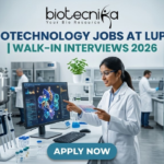 Biotechnology Jobs at Lupin | Walk-In Interviews 2026. A high-quality, 1280x720 featured image for "Biotechnology Jobs at Lupin | Walk-In Interviews 2026." The image features the Biotecnika logo at the top center. In a modern, bright industrial laboratory, a female scientist in a white lab coat and safety goggles points to a monitor displaying a detailed 3D protein structure. Large stainless steel bioreactors and advanced lab equipment are visible in the background, creating a realistic pharmaceutical R&D atmosphere. The main title is displayed in bold teal and dark blue lettering, and a prominent blue "APPLY NOW" button is positioned at the bottom.