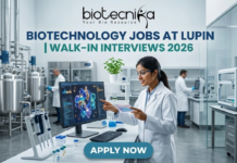 Lupin Walk-In Interviews 2026 | Biotechnology Jobs in R&D, Manufacturing & Quality Biotechnology Jobs at Lupin | Walk-In Interviews 2026. A high-quality, 1280x720 featured image for "Biotechnology Jobs at Lupin | Walk-In Interviews 2026." The image features the Biotecnika logo at the top center. In a modern, bright industrial laboratory, a female scientist in a white lab coat and safety goggles points to a monitor displaying a detailed 3D protein structure. Large stainless steel bioreactors and advanced lab equipment are visible in the background, creating a realistic pharmaceutical R&D atmosphere. The main title is displayed in bold teal and dark blue lettering, and a prominent blue "APPLY NOW" button is positioned at the bottom.