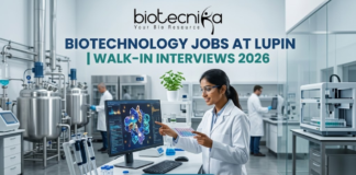 Lupin Walk-In Interviews 2026 | Biotechnology Jobs in R&D, Manufacturing & Quality Biotechnology Jobs at Lupin | Walk-In Interviews 2026. A high-quality, 1280x720 featured image for "Biotechnology Jobs at Lupin | Walk-In Interviews 2026." The image features the Biotecnika logo at the top center. In a modern, bright industrial laboratory, a female scientist in a white lab coat and safety goggles points to a monitor displaying a detailed 3D protein structure. Large stainless steel bioreactors and advanced lab equipment are visible in the background, creating a realistic pharmaceutical R&D atmosphere. The main title is displayed in bold teal and dark blue lettering, and a prominent blue "APPLY NOW" button is positioned at the bottom.