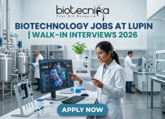 Lupin Walk-In Interviews 2026 | Biotechnology Jobs in R&D, Manufacturing & Quality Biotechnology Jobs at Lupin | Walk-In Interviews 2026. A high-quality, 1280x720 featured image for "Biotechnology Jobs at Lupin | Walk-In Interviews 2026." The image features the Biotecnika logo at the top center. In a modern, bright industrial laboratory, a female scientist in a white lab coat and safety goggles points to a monitor displaying a detailed 3D protein structure. Large stainless steel bioreactors and advanced lab equipment are visible in the background, creating a realistic pharmaceutical R&D atmosphere. The main title is displayed in bold teal and dark blue lettering, and a prominent blue "APPLY NOW" button is positioned at the bottom.