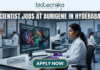 High-Paying Scientist Jobs in Hyderabad – Aurigene Hiring MSc Life Science Graduates Now! Scientist Jobs at Aurigene in Hyderabad. A high-quality, professional featured image for "Scientist Jobs at Aurigene in Hyderabad." The scene depicts a female scientist in a white lab coat and purple gloves working in a sophisticated laboratory. She is focused on a large monitor displaying vibrant 3D molecular structures under the heading "Drug Discovery Analysis." Her desk features a modern microscope and a small plaque that says "Workplace: Hyderabad Science Hub." In the background, the iconic Charminar is visible through a large window, establishing the Hyderabad setting. The Biotecnika logo is positioned at the top center, the job title is in bold white lettering, and a prominent "APPLY NOW" button is at the bottom.