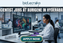 High-Paying Scientist Jobs in Hyderabad – Aurigene Hiring MSc Life Science Graduates Now! Scientist Jobs at Aurigene in Hyderabad. A high-quality, professional featured image for "Scientist Jobs at Aurigene in Hyderabad." The scene depicts a female scientist in a white lab coat and purple gloves working in a sophisticated laboratory. She is focused on a large monitor displaying vibrant 3D molecular structures under the heading "Drug Discovery Analysis." Her desk features a modern microscope and a small plaque that says "Workplace: Hyderabad Science Hub." In the background, the iconic Charminar is visible through a large window, establishing the Hyderabad setting. The Biotecnika logo is positioned at the top center, the job title is in bold white lettering, and a prominent "APPLY NOW" button is at the bottom.