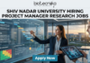 Research Jobs at Shiv Nadar University | Apply Now. A high-quality, realistic featured image for a "Project Manager Research Jobs" opening at Shiv Nadar University. The scene features a professional woman in business attire interacting with a large, glowing transparent digital dashboard that displays research project timelines (Gantt charts), risk assessment matrices, and milestones for "Advanced Cell Therapy." A senior male colleague stands nearby holding a tablet with a 3D molecular visualization. The setting is a sleek, modern office with large windows overlooking a university campus at sunset. The Biotecnika logo is at the top, the main job title is in bold black text, and a prominent blue "Apply Now" button is at the bottom.