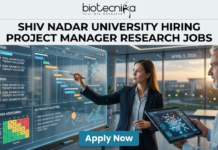 Research Jobs At Shiv Nadar University | Earn Upto ₹42,000/Month | Apply Now! Research Jobs at Shiv Nadar University | Apply Now. A high-quality, realistic featured image for a "Project Manager Research Jobs" opening at Shiv Nadar University. The scene features a professional woman in business attire interacting with a large, glowing transparent digital dashboard that displays research project timelines (Gantt charts), risk assessment matrices, and milestones for "Advanced Cell Therapy." A senior male colleague stands nearby holding a tablet with a 3D molecular visualization. The setting is a sleek, modern office with large windows overlooking a university campus at sunset. The Biotecnika logo is at the top, the main job title is in bold black text, and a prominent blue "Apply Now" button is at the bottom.
