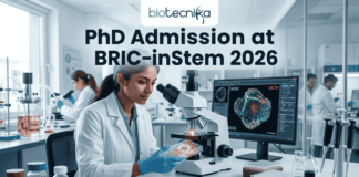 PhD Admission at BRIC-inStem 2026 | JRF-Based Entry + Cutting-Edge Stem Cell Research Opportunity! A high-resolution featured image for PhD Admission at BRIC-inStem 2026. The image depicts a modern, realistic biotechnology laboratory where a female scientist of Indian descent, wearing a lab coat and safety goggles, is carefully examining a multi-well plate. Beside her, a computer monitor displays a vibrant, high-detail microscopic image of a stem cell labeled with genetic markers. The Biotecnika logo is positioned at the top center, and a prominent "APPLY NOW" button is located at the bottom.