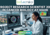 Project Research Scientist Job in Cancer Biology at MAHE. A professional featured image for a job posting. At the center, a female scientist in a white lab coat smiles while working with a multi-channel pipette in a high-tech laboratory. Behind her, large computer monitors display vibrant 3D visualizations of cancer cells and molecular data. A window in the background offers a scenic view of a coastal campus. At the top center is the Biotecnika logo. Large, bold white text across the upper half reads: "PROJECT RESEARCH SCIENTIST JOB IN CANCER BIOLOGY AT MAHE". At the bottom center, there is a prominent, glowing green and blue "APPLY NOW" button.