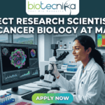 Project Research Scientist Job in Cancer Biology at MAHE. A professional featured image for a job posting. At the center, a female scientist in a white lab coat smiles while working with a multi-channel pipette in a high-tech laboratory. Behind her, large computer monitors display vibrant 3D visualizations of cancer cells and molecular data. A window in the background offers a scenic view of a coastal campus. At the top center is the Biotecnika logo. Large, bold white text across the upper half reads: "PROJECT RESEARCH SCIENTIST JOB IN CANCER BIOLOGY AT MAHE". At the bottom center, there is a prominent, glowing green and blue "APPLY NOW" button.