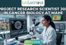 Project Research Scientist Job in Cancer Biology – MAHE Hiring Now with ₹61K Salary! Project Research Scientist Job in Cancer Biology at MAHE. A professional featured image for a job posting. At the center, a female scientist in a white lab coat smiles while working with a multi-channel pipette in a high-tech laboratory. Behind her, large computer monitors display vibrant 3D visualizations of cancer cells and molecular data. A window in the background offers a scenic view of a coastal campus. At the top center is the Biotecnika logo. Large, bold white text across the upper half reads: "PROJECT RESEARCH SCIENTIST JOB IN CANCER BIOLOGY AT MAHE". At the bottom center, there is a prominent, glowing green and blue "APPLY NOW" button.