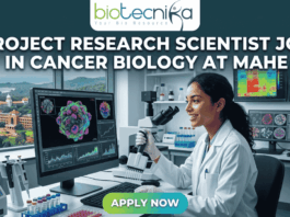 Project Research Scientist Job in Cancer Biology at MAHE. A professional featured image for a job posting. At the center, a female scientist in a white lab coat smiles while working with a multi-channel pipette in a high-tech laboratory. Behind her, large computer monitors display vibrant 3D visualizations of cancer cells and molecular data. A window in the background offers a scenic view of a coastal campus. At the top center is the Biotecnika logo. Large, bold white text across the upper half reads: "PROJECT RESEARCH SCIENTIST JOB IN CANCER BIOLOGY AT MAHE". At the bottom center, there is a prominent, glowing green and blue "APPLY NOW" button.