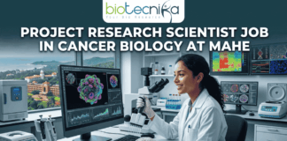 Project Research Scientist Job in Cancer Biology at MAHE. A professional featured image for a job posting. At the center, a female scientist in a white lab coat smiles while working with a multi-channel pipette in a high-tech laboratory. Behind her, large computer monitors display vibrant 3D visualizations of cancer cells and molecular data. A window in the background offers a scenic view of a coastal campus. At the top center is the Biotecnika logo. Large, bold white text across the upper half reads: "PROJECT RESEARCH SCIENTIST JOB IN CANCER BIOLOGY AT MAHE". At the bottom center, there is a prominent, glowing green and blue "APPLY NOW" button.