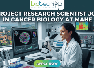 Project Research Scientist Job in Cancer Biology – MAHE Hiring Now with ₹61K Salary! Project Research Scientist Job in Cancer Biology at MAHE. A professional featured image for a job posting. At the center, a female scientist in a white lab coat smiles while working with a multi-channel pipette in a high-tech laboratory. Behind her, large computer monitors display vibrant 3D visualizations of cancer cells and molecular data. A window in the background offers a scenic view of a coastal campus. At the top center is the Biotecnika logo. Large, bold white text across the upper half reads: "PROJECT RESEARCH SCIENTIST JOB IN CANCER BIOLOGY AT MAHE". At the bottom center, there is a prominent, glowing green and blue "APPLY NOW" button.