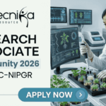 Research Associate Job At BRIC-NIPGR | Life Science Opportunity. A professional featured image designed for a job page. On the left side, the Biotecnika logo is at the top, followed by the bold text "RESEARCH ASSOCIATE Opportunity 2026 at BRIC-NIPGR". On the right side, a realistic image depicts a female scientist in a white lab coat and safety glasses working in a modern, high-tech plant research laboratory; she is using a pipette to study small green seedlings. The background features monitors displaying scientific plant data. At the bottom center, there is a prominent blue "APPLY NOW" button.