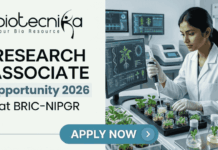Research Associate Opportunity 2026 at BRIC-NIPGR | Earn Upto ₹58,000/month + HRA Research Associate Job At BRIC-NIPGR | Life Science Opportunity. A professional featured image designed for a job page. On the left side, the Biotecnika logo is at the top, followed by the bold text "RESEARCH ASSOCIATE Opportunity 2026 at BRIC-NIPGR". On the right side, a realistic image depicts a female scientist in a white lab coat and safety glasses working in a modern, high-tech plant research laboratory; she is using a pipette to study small green seedlings. The background features monitors displaying scientific plant data. At the bottom center, there is a prominent blue "APPLY NOW" button.