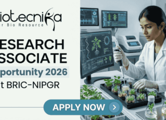 Research Associate Job At BRIC-NIPGR | Life Science Opportunity. A professional featured image designed for a job page. On the left side, the Biotecnika logo is at the top, followed by the bold text "RESEARCH ASSOCIATE Opportunity 2026 at BRIC-NIPGR". On the right side, a realistic image depicts a female scientist in a white lab coat and safety glasses working in a modern, high-tech plant research laboratory; she is using a pipette to study small green seedlings. The background features monitors displaying scientific plant data. At the bottom center, there is a prominent blue "APPLY NOW" button.