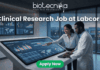 Clinical Research Job at Labcorp Bangalore | Apply Now! A realistic and attention-grabbing featured image, size 1280x720, designed for a job page. It features two black clinical researchers, a woman pointing and a man in a white lab coat, collaborating intensely at a massive, tilted touch-screen interface within a futuristic laboratory setting. The screen displays complex multi-colored data visualizations, timelines, maps, and charts with text labeling "Clinical Trial Data Analysis". Prominent white text at the top states: "Clinical Research Job at Labcorp". The complete Biotecnika logo (including text and tagline 'Your Bio Resource') is at the top-center. A distinct vibrant green "Apply Now" button is clearly positioned at the bottom-center.