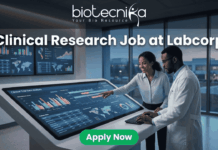 Clinical Research Job at Labcorp Bangalore | Apply Now! A realistic and attention-grabbing featured image, size 1280x720, designed for a job page. It features two black clinical researchers, a woman pointing and a man in a white lab coat, collaborating intensely at a massive, tilted touch-screen interface within a futuristic laboratory setting. The screen displays complex multi-colored data visualizations, timelines, maps, and charts with text labeling "Clinical Trial Data Analysis". Prominent white text at the top states: "Clinical Research Job at Labcorp". The complete Biotecnika logo (including text and tagline 'Your Bio Resource') is at the top-center. A distinct vibrant green "Apply Now" button is clearly positioned at the bottom-center.