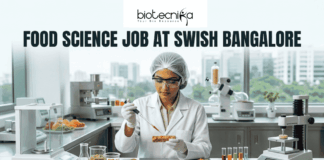 Food Science Job at Swish Bangalore. A clean, modern featured image showing a food scientist in a sterile lab environment using a precision pipette on a nutrient-rich food prototype. The lab is equipped with advanced food testing machinery and colorful test tubes. The city skyline of Bangalore is visible through the window. The Biotecnika logo is at the top, and an orange "Apply Now" button is at the bottom.