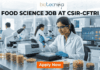 Food Science Job at CSIR-CFTRI Earn Upto ₹31,000 + HRA. A professional recruitment banner for a Food Science job at CSIR-CFTRI. The image features a female scientist in a white lab coat and safety glasses working in a modern, brightly lit laboratory. She is using a pipette to analyze samples in a multi-well plate. The foreground displays glass jars filled with various grains and food ingredients. The Biotecnika logo is at the top center, and the main text reads "FOOD SCIENCE JOB AT CSIR-CFTRI" in bold black letters. A prominent orange "Apply Now" button is positioned at the bottom.