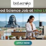 Food Science Job at Isha LIFE R&D Specialist Role. A featured image for a job posting titled "Food Science Job at Isha LIFE." The image displays a professional female scientist in a white lab coat working in a modern, high-tech food laboratory. On the lab counter, there are various bowls of natural grains, herbs, and scientific testing equipment. Through the large glass windows in the background, the iconic Adi Yogi statue is visible against a scenic mountain landscape, reflecting the Isha Foundation theme. The Biotecnika logo is positioned at the top center, and a prominent green "APPLY NOW" button is located at the bottom.