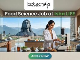 Food Science Job at Isha LIFE R&D Specialist Role. A featured image for a job posting titled "Food Science Job at Isha LIFE." The image displays a professional female scientist in a white lab coat working in a modern, high-tech food laboratory. On the lab counter, there are various bowls of natural grains, herbs, and scientific testing equipment. Through the large glass windows in the background, the iconic Adi Yogi statue is visible against a scenic mountain landscape, reflecting the Isha Foundation theme. The Biotecnika logo is positioned at the top center, and a prominent green "APPLY NOW" button is located at the bottom.
