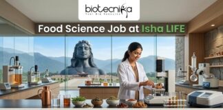 Food Science Job at Isha LIFE R&D Specialist Role. A featured image for a job posting titled "Food Science Job at Isha LIFE." The image displays a professional female scientist in a white lab coat working in a modern, high-tech food laboratory. On the lab counter, there are various bowls of natural grains, herbs, and scientific testing equipment. Through the large glass windows in the background, the iconic Adi Yogi statue is visible against a scenic mountain landscape, reflecting the Isha Foundation theme. The Biotecnika logo is positioned at the top center, and a prominent green "APPLY NOW" button is located at the bottom.