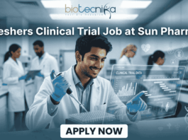 Freshers Clinical Trial Job at Sun Pharma | Don't Miss It. A featured image for a job posting titled "Freshers Clinical Trial Job at Sun Pharma" in a $1280 \times 720$ format.The image depicts a modern, high-tech laboratory setting where a young, smiling male scientist in a white lab coat is working with a pipette and a multi-well plate. A transparent digital holographic display in front of him shows "Clinical Trial Data" with various charts and graphs. In the background, other researchers are visible in a clean, professional clinical environment.The Biotecnika logo is positioned clearly at the top center, and a prominent, rounded "APPLY NOW" button is located at the bottom center. The overall aesthetic is professional, bright, and engaging.