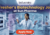 Fresher's Job Alert! Biotechnology Job at Sun Pharma Don’t Miss Out! A promotional job banner with the Biotecnika logo at the top. The headline text is "Fresher's Biotechnology Job" and "at Sun Pharma". A smiling young woman in a lab coat stands in a futuristic laboratory, interacting with floating holographic screens. The screens display molecular structures labeled "Drug Discovery" and "Lead Identification," and a DNA helix with graphs labeled "DNA" and "Data Discovery". A large red button at the bottom says "Apply Now". The background shows complex lab equipment.