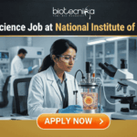 Govt Life Science Job Alert 2026 at National Institute of Biologicals. A professional featured image for a "Govt Life Science Job at National Institute of Biologicals." It shows a scientist in a clean, modern laboratory setting operating a bioreactor. The design features a prominent Biotecnika logo at the top, the Indian national emblem (Satyameva Jayate) in a frame on the desk, and a bright orange "Apply Now" button at the bottom.