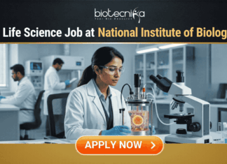 Govt. Life Science Job Alert 2026 at National Institute of Biologicals | Apply Now Govt Life Science Job Alert 2026 at National Institute of Biologicals. A professional featured image for a "Govt Life Science Job at National Institute of Biologicals." It shows a scientist in a clean, modern laboratory setting operating a bioreactor. The design features a prominent Biotecnika logo at the top, the Indian national emblem (Satyameva Jayate) in a frame on the desk, and a bright orange "Apply Now" button at the bottom.