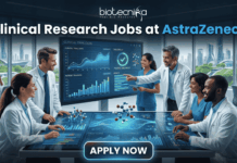 Hiring Now! Clinical Research Jobs at AstraZeneca for Life Science Graduates! A professional featured image for "Clinical Research Jobs at AstraZeneca." It shows a group of medical researchers in lab coats gathered around a large interactive touch-table displaying clinical trial data, graphs, and 3D molecular models. The Biotecnika logo is at the top, and a sleek "Apply Now" button is at the bottom.
