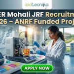 Junior Research Fellow (JRF) at IISER Mohali | Apply Now. A professional promotional image for the IISER Mohali JRF Recruitment 2026. At the top, the Biotecnika logo is displayed above the bold headline: "IISER Mohali JRF Recruitment 2026 – ANRF Funded Project." The scene features a young Indian female scientist in a white lab coat and safety glasses, meticulously working with a sophisticated bioreactor containing an orange-tinted culture. The background shows a bright, modern research facility with other scientists at work and a large window overlooking a lush, green university campus. To the left, a whiteboard is covered in complex chemical equations and the words "JRF PROJECT 2026." A prominent, glowing green "APPLY NOW" button is centered at the bottom of the frame.