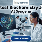 Latest Biochemistry Job At Syngene Don't Miss This Opportunity. A realistic and professional featured image for a biochemistry job vacancy at Syngene. The image shows a female scientist in a white lab coat working in a state-of-the-art laboratory, focusing on a colorful 3D molecular protein structure displayed on a large computer screen. The Biotecnika logo is at the top, the headline "Latest Biochemistry Job At Syngene" is prominently displayed, and a blue "Apply Now" button is positioned at the bottom center.