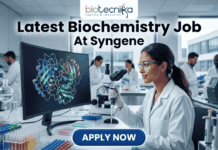 Latest Biochemistry Job At Syngene | Apply For Associate Scientist Role Today! Latest Biochemistry Job At Syngene Don't Miss This Opportunity. A realistic and professional featured image for a biochemistry job vacancy at Syngene. The image shows a female scientist in a white lab coat working in a state-of-the-art laboratory, focusing on a colorful 3D molecular protein structure displayed on a large computer screen. The Biotecnika logo is at the top, the headline "Latest Biochemistry Job At Syngene" is prominently displayed, and a blue "Apply Now" button is positioned at the bottom center.