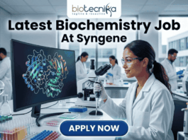 Latest Biochemistry Job At Syngene Don't Miss This Opportunity. A realistic and professional featured image for a biochemistry job vacancy at Syngene. The image shows a female scientist in a white lab coat working in a state-of-the-art laboratory, focusing on a colorful 3D molecular protein structure displayed on a large computer screen. The Biotecnika logo is at the top, the headline "Latest Biochemistry Job At Syngene" is prominently displayed, and a blue "Apply Now" button is positioned at the bottom center.