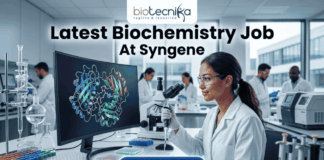 Latest Biochemistry Job At Syngene Don't Miss This Opportunity. A realistic and professional featured image for a biochemistry job vacancy at Syngene. The image shows a female scientist in a white lab coat working in a state-of-the-art laboratory, focusing on a colorful 3D molecular protein structure displayed on a large computer screen. The Biotecnika logo is at the top, the headline "Latest Biochemistry Job At Syngene" is prominently displayed, and a blue "Apply Now" button is positioned at the bottom center.