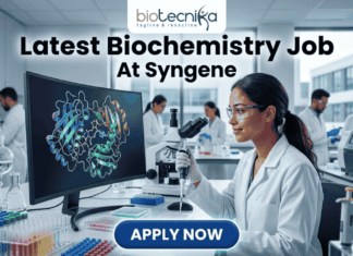 Latest Biochemistry Job At Syngene | Apply For Associate Scientist Role Today! Latest Biochemistry Job At Syngene Don't Miss This Opportunity. A realistic and professional featured image for a biochemistry job vacancy at Syngene. The image shows a female scientist in a white lab coat working in a state-of-the-art laboratory, focusing on a colorful 3D molecular protein structure displayed on a large computer screen. The Biotecnika logo is at the top, the headline "Latest Biochemistry Job At Syngene" is prominently displayed, and a blue "Apply Now" button is positioned at the bottom center.