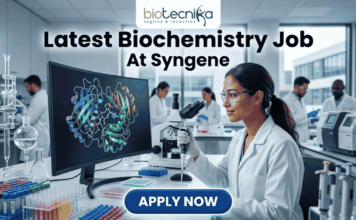 Latest Biochemistry Job At Syngene | Apply For Associate Scientist Role Today! Latest Biochemistry Job At Syngene Don't Miss This Opportunity. A realistic and professional featured image for a biochemistry job vacancy at Syngene. The image shows a female scientist in a white lab coat working in a state-of-the-art laboratory, focusing on a colorful 3D molecular protein structure displayed on a large computer screen. The Biotecnika logo is at the top, the headline "Latest Biochemistry Job At Syngene" is prominently displayed, and a blue "Apply Now" button is positioned at the bottom center.