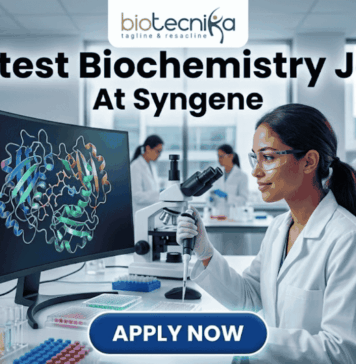Latest Biochemistry Job At Syngene | Apply For Associate Scientist Role Today! Latest Biochemistry Job At Syngene Don't Miss This Opportunity. A realistic and professional featured image for a biochemistry job vacancy at Syngene. The image shows a female scientist in a white lab coat working in a state-of-the-art laboratory, focusing on a colorful 3D molecular protein structure displayed on a large computer screen. The Biotecnika logo is at the top, the headline "Latest Biochemistry Job At Syngene" is prominently displayed, and a blue "Apply Now" button is positioned at the bottom center.