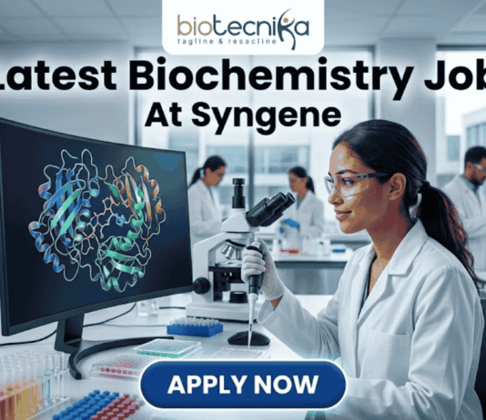 Latest Biochemistry Job At Syngene Don't Miss This Opportunity. A realistic and professional featured image for a biochemistry job vacancy at Syngene. The image shows a female scientist in a white lab coat working in a state-of-the-art laboratory, focusing on a colorful 3D molecular protein structure displayed on a large computer screen. The Biotecnika logo is at the top, the headline "Latest Biochemistry Job At Syngene" is prominently displayed, and a blue "Apply Now" button is positioned at the bottom center.
