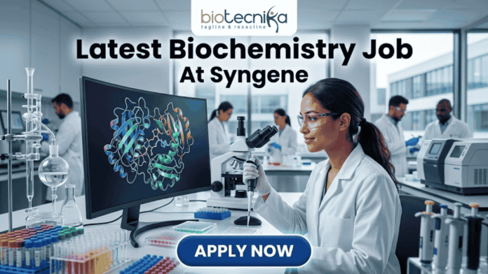 Latest Biochemistry Job At Syngene Don't Miss This Opportunity Latest Biochemistry Job At Syngene Don't Miss This Opportunity. A realistic and professional featured image for a biochemistry job vacancy at Syngene. The image shows a female scientist in a white lab coat working in a state-of-the-art laboratory, focusing on a colorful 3D molecular protein structure displayed on a large computer screen. The Biotecnika logo is at the top, the headline "Latest Biochemistry Job At Syngene" is prominently displayed, and a blue "Apply Now" button is positioned at the bottom center.