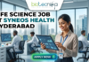 Life Science Job at Syneos Health Hyderabad. A career advertisement banner for a life science job at Syneos Health in Hyderabad, presented by Biotecnika Global. The banner features a smiling woman in a lab coat and safety glasses, holding and pointing at a tablet that displays data charts and a molecular structure. She is standing in a modern office and laboratory setting, with large windows overlooking a city skyline. Other researchers in lab coats are seated at desks in the background, working on computer monitors. The "biotecnika Global Hiring and Neurnic Solutions" logo is at the top, and a green "Apply now" button with an arrow is at the bottom.