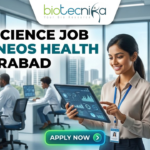 Life Science Job at Syneos Health Hyderabad. A career advertisement banner for a life science job at Syneos Health in Hyderabad, presented by Biotecnika Global. The banner features a smiling woman in a lab coat and safety glasses, holding and pointing at a tablet that displays data charts and a molecular structure. She is standing in a modern office and laboratory setting, with large windows overlooking a city skyline. Other researchers in lab coats are seated at desks in the background, working on computer monitors. The "biotecnika Global Hiring and Neurnic Solutions" logo is at the top, and a green "Apply now" button with an arrow is at the bottom.