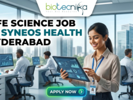 Life Science Job at Syneos Health Hyderabad. A career advertisement banner for a life science job at Syneos Health in Hyderabad, presented by Biotecnika Global. The banner features a smiling woman in a lab coat and safety glasses, holding and pointing at a tablet that displays data charts and a molecular structure. She is standing in a modern office and laboratory setting, with large windows overlooking a city skyline. Other researchers in lab coats are seated at desks in the background, working on computer monitors. The "biotecnika Global Hiring and Neurnic Solutions" logo is at the top, and a green "Apply now" button with an arrow is at the bottom.