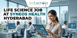 Life Science Job at Syneos Health Hyderabad. A career advertisement banner for a life science job at Syneos Health in Hyderabad, presented by Biotecnika Global. The banner features a smiling woman in a lab coat and safety glasses, holding and pointing at a tablet that displays data charts and a molecular structure. She is standing in a modern office and laboratory setting, with large windows overlooking a city skyline. Other researchers in lab coats are seated at desks in the background, working on computer monitors. The "biotecnika Global Hiring and Neurnic Solutions" logo is at the top, and a green "Apply now" button with an arrow is at the bottom.