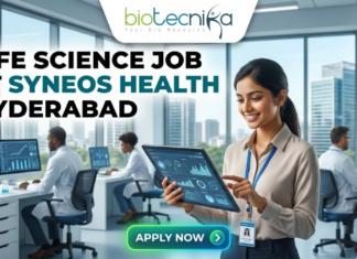 Exciting Life Science Job at Syneos Health Hyderabad | BSc/MSc Candidates Apply Now Life Science Job at Syneos Health Hyderabad. A career advertisement banner for a life science job at Syneos Health in Hyderabad, presented by Biotecnika Global. The banner features a smiling woman in a lab coat and safety glasses, holding and pointing at a tablet that displays data charts and a molecular structure. She is standing in a modern office and laboratory setting, with large windows overlooking a city skyline. Other researchers in lab coats are seated at desks in the background, working on computer monitors. The "biotecnika Global Hiring and Neurnic Solutions" logo is at the top, and a green "Apply now" button with an arrow is at the bottom.