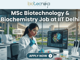 MSc Biotechnology & Biochemistry Job at IIT Delhi. A professional and realistic featured image for a job opening. At the top center is the Biotecnika Global logo, followed by large, bold text reading "MSc Biotechnology & Biochemistry Job at IIT Delhi." The central scene depicts a female researcher in a white lab coat working in a high-tech laboratory, using a pipette on colorful samples while viewing a 3D molecular structure on a large monitor. The background shows advanced lab equipment and a clear view of the IIT Delhi Research campus through large windows. At the bottom, there is a prominent teal button that says "APPLY NOW" with a white arrow icon.